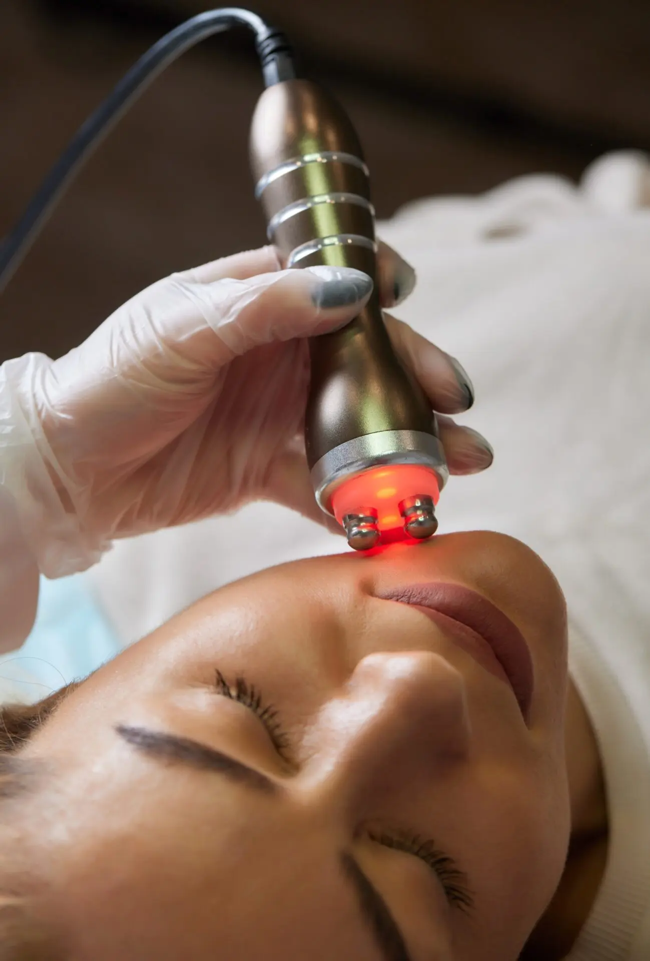 A cosmetologist in rubber gloves does the rf-lifting procedure on the face. In the background, the Rf cosmetology device. Close up. Side view. The concept of professional cosmetology and rejuvenation.<br />
